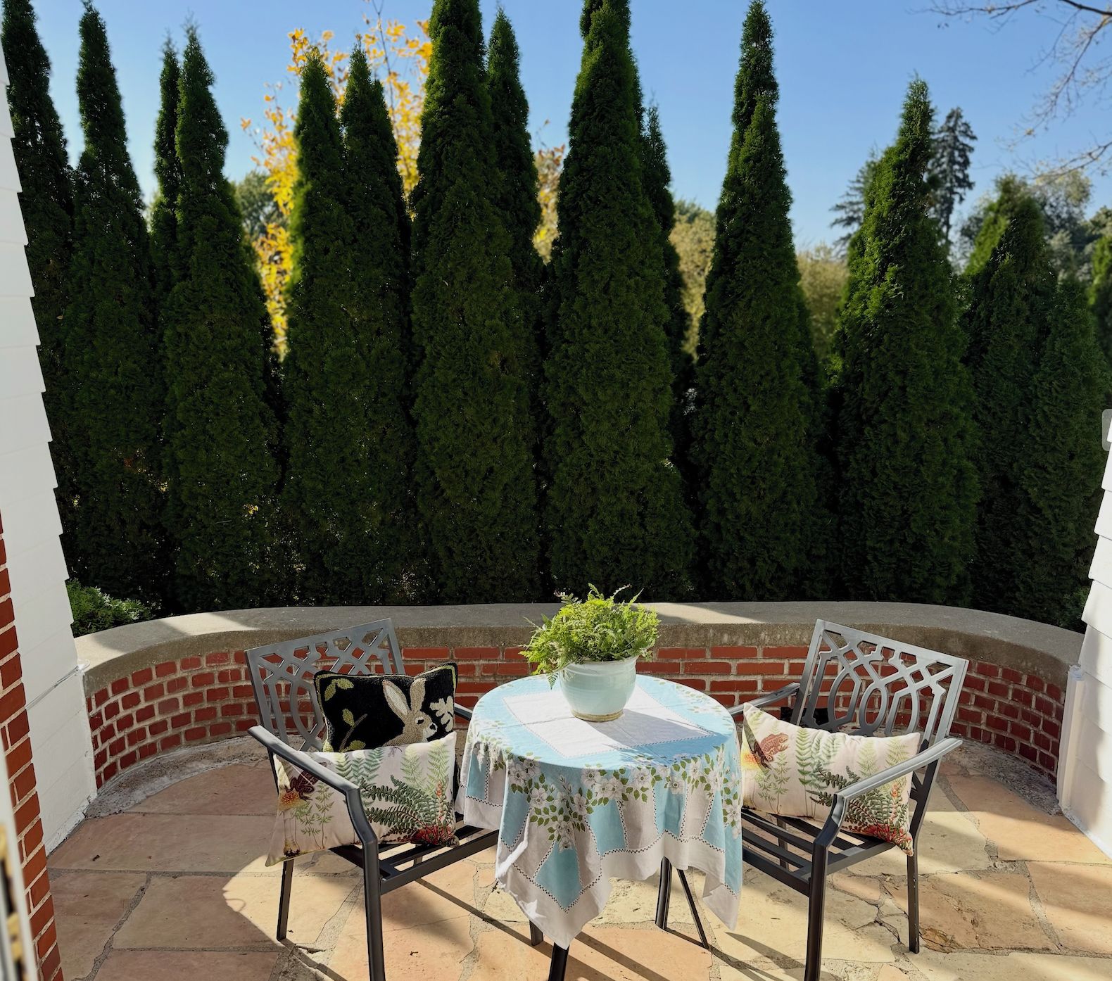 An outdoor patio with a round table and chairs, surrounded by a brick wall and tall trees.