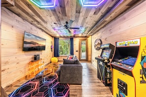 A room with wooden walls, a ceiling fan, a television, a couch, a gaming console, and a window with curtains. - Indoor Pool • Arcade • Hot Tub  Pigeon Forge (Pigeon Forge)