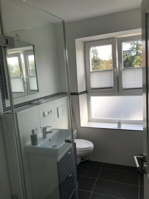 A bathroom with a glass shower enclosure, a white sink with a faucet, a toilet, and a window with blinds. - New Listing (Altenkrempe)