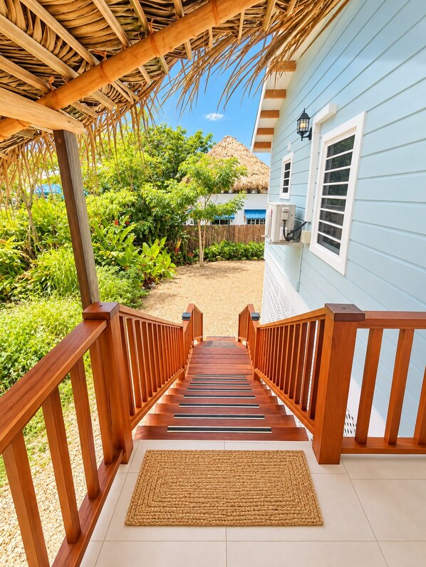 A wooden staircase leading to a house with a thatched roof, surrounded by greenery.