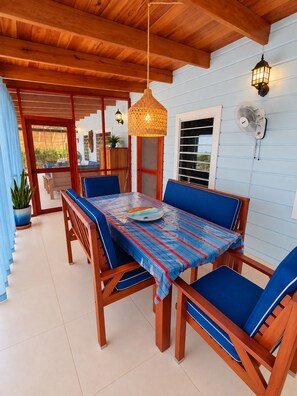 A covered outdoor dining area with a wooden table and blue cushioned chairs, a hanging lantern, and a wall-mounted fan.