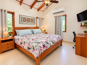 A bedroom with a bed, bedside tables, a ceiling fan, an air conditioning unit, a TV, and a desk with a chair.