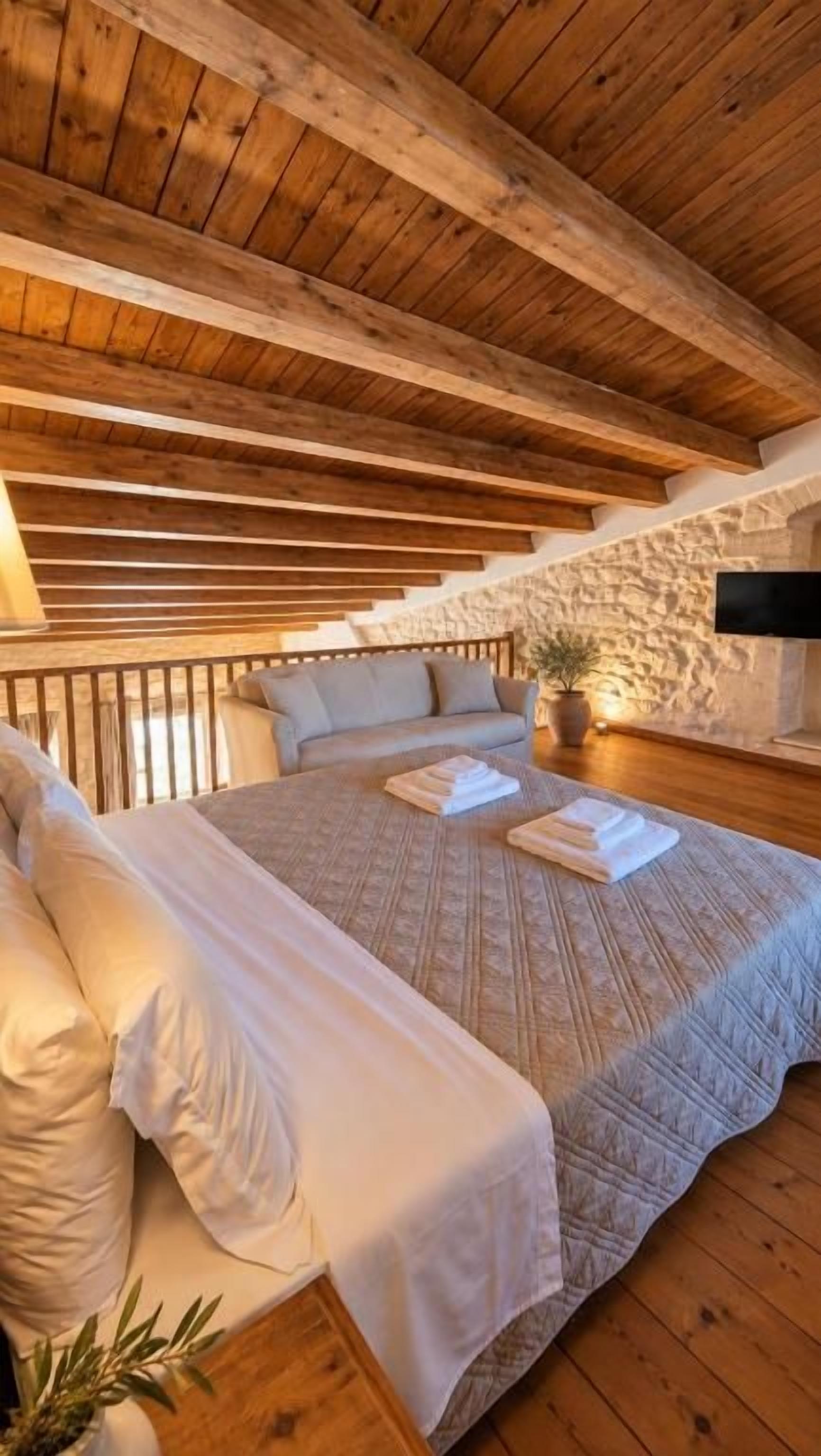 Une chambre avec un plafond en bois, un lit recouvert de draps blancs, un mur en pierre et un téléviseur à écran plat fixé au mur.
