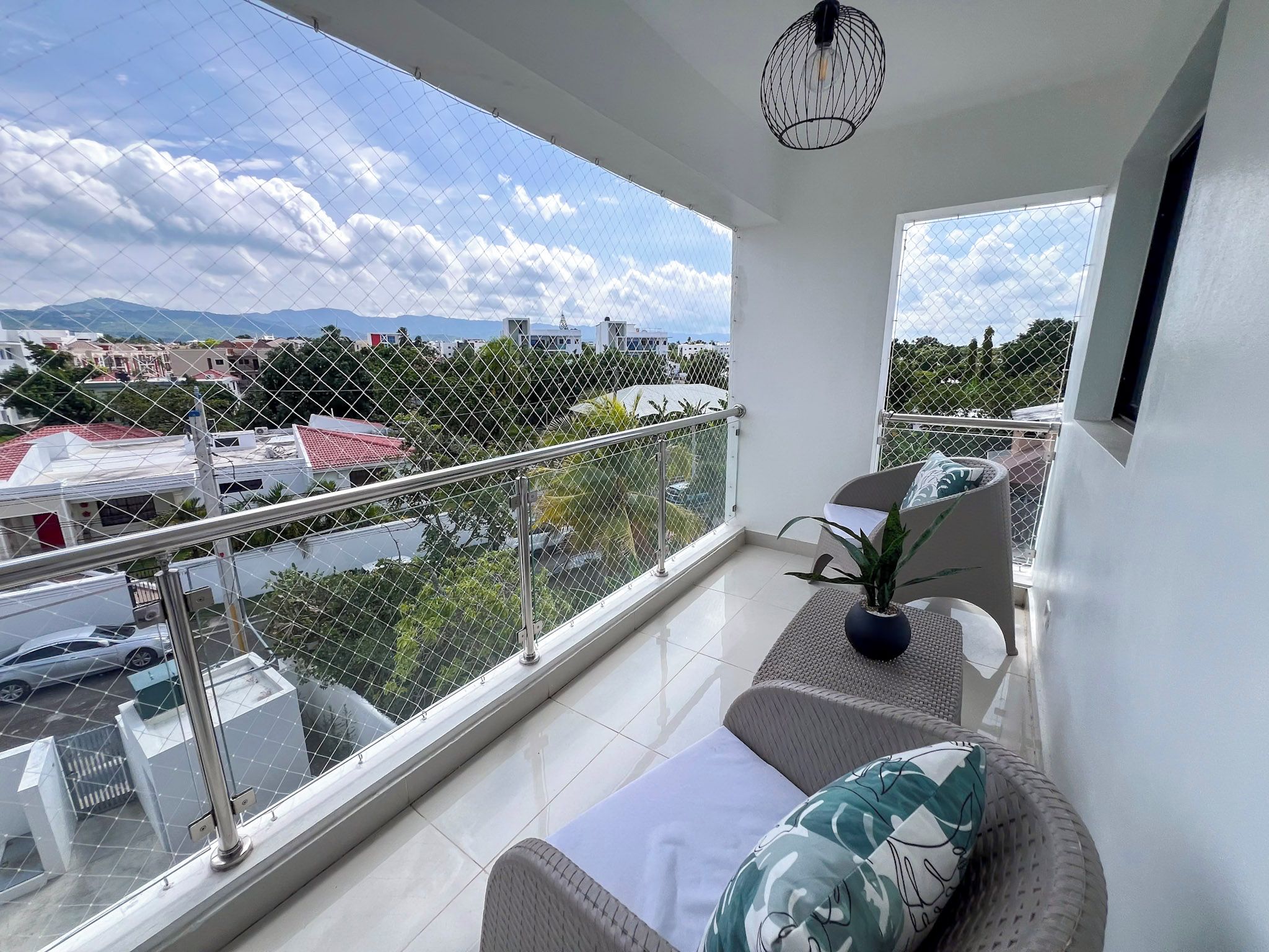 A balcony with a view of a cityscape, a potted plant, and a seating area with cushions.