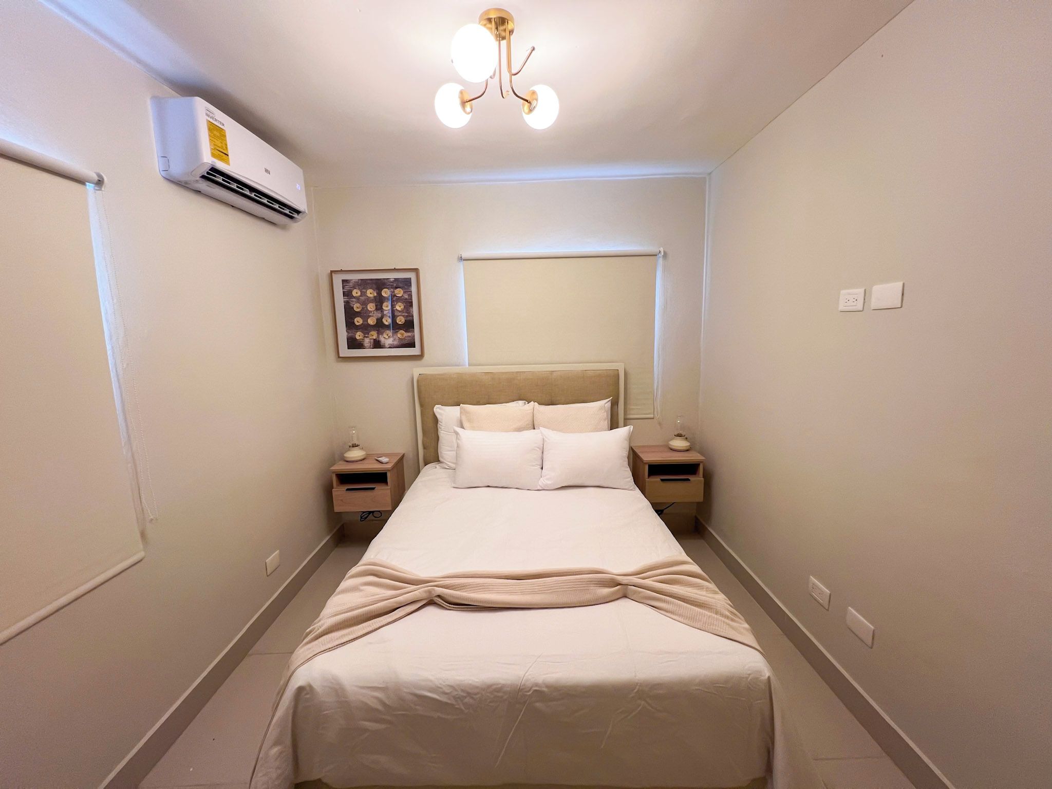 A hotel room with a bed, bedside tables, an air conditioning unit, a wall-mounted picture, and a ceiling light fixture.