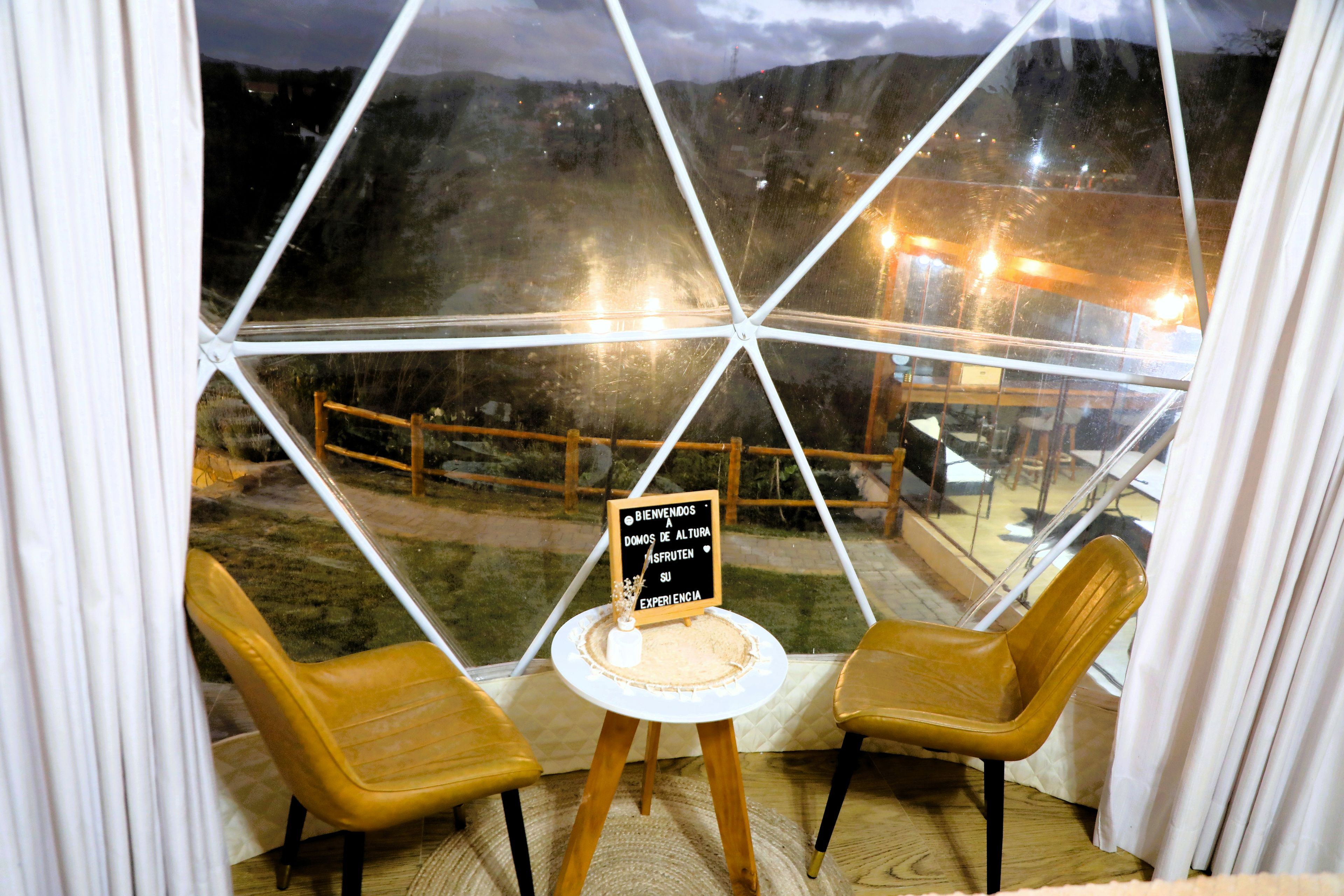 Una habitación con una estructura de cúpula de cristal, dos sillas, una mesita y un cartel con texto.