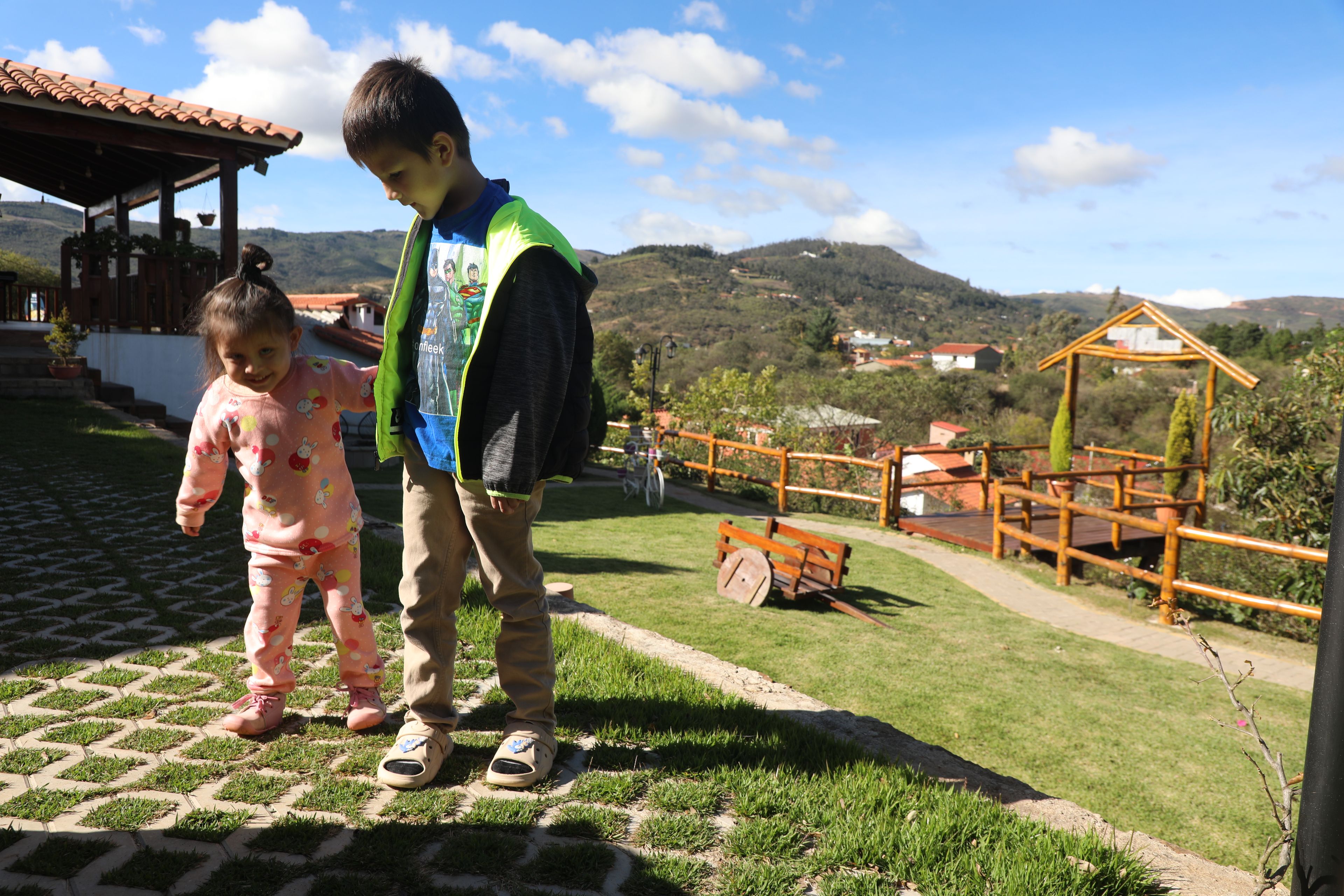 Un niño y un bebé están de pie en una zona pavimentada, con un campo de césped y estructuras de madera al fondo.
