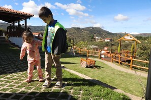 A child and a toddler standing on a paved area with a grassy field and wooden structures in the background.
