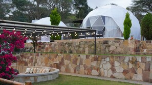 A stone wall with a seating area, a pergola with string lights, and a dome-shaped structure in the background.