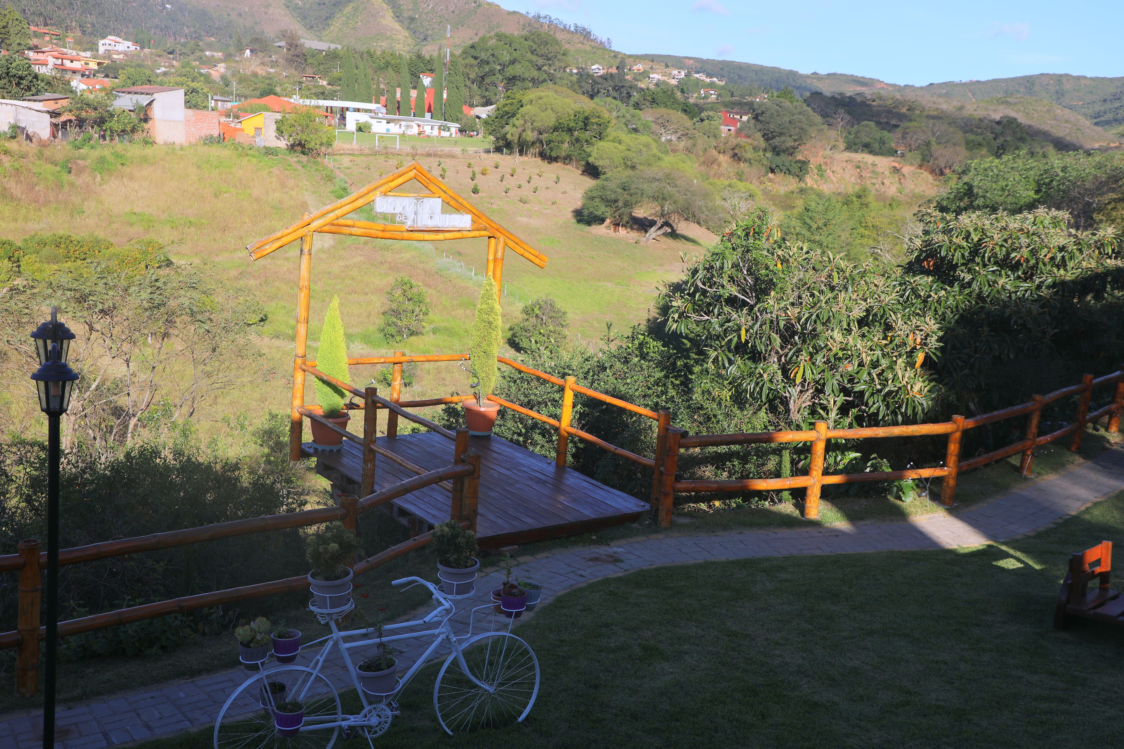 Una terraza de madera con una bicicleta, rodeada de follaje y vistas a una colina con casas.