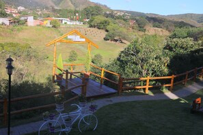A wooden deck with a bicycle, surrounded by greenery and a view of a hillside with houses.