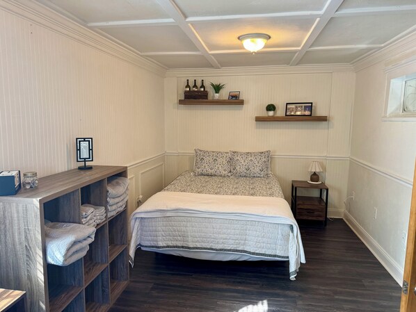 Un dormitorio con una cama, una mesita de noche, un estante con botellas y una pequeña planta.
