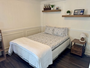 Una cama bien hecha con una colcha estampada, una mesita de noche con lámpara y dos repisas en la pared con objetos decorativos.