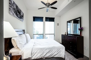 Une chambre à coucher comprenant un lit, une commode, un miroir, un ventilateur de plafond et une fenêtre avec des stores.