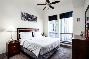 Une chambre d’hôtel équipée d’un lit, d’une table de chevet, d’une lampe, d’une horloge numérique, d’un ventilateur de plafond et d’une grande fenêtre donnant sur la ville.