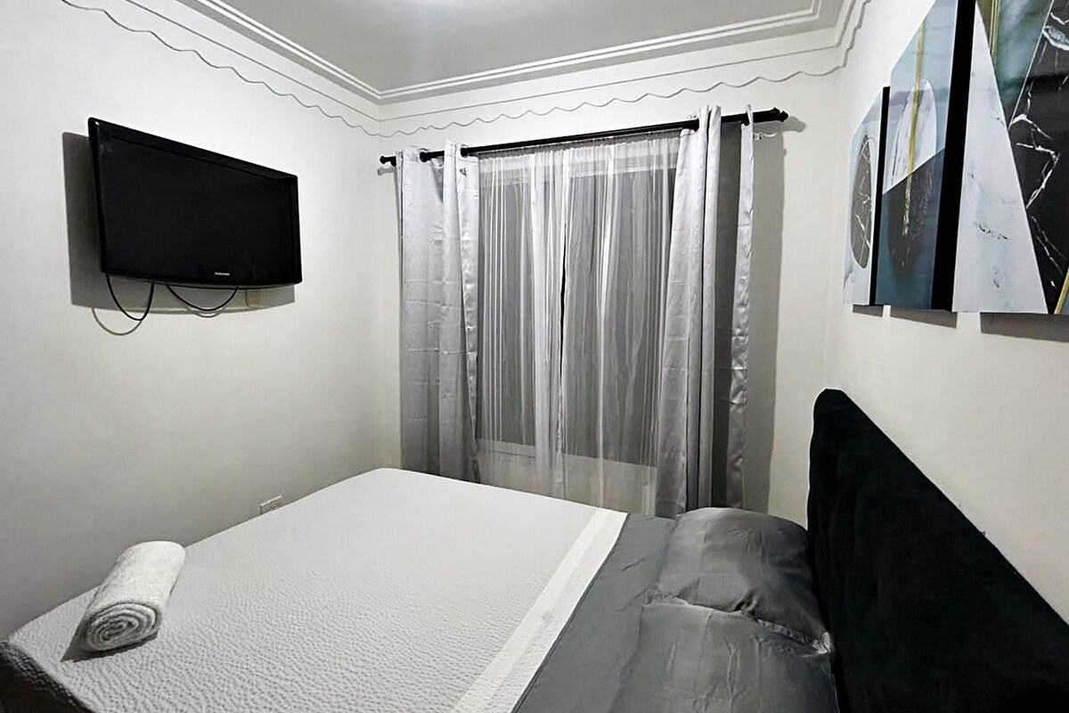 A bedroom with a bed, a television mounted on the wall, and a window with sheer curtains.