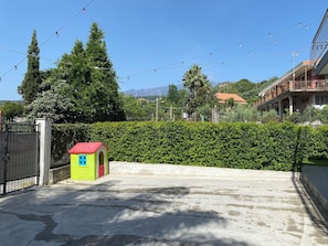 Ein begrenzter Eingangsbereich mit einer kleinen, grün-rot gestreiften Struktur, einer Hecke und einem Gebäude mit rotem Dach im Hintergrund.