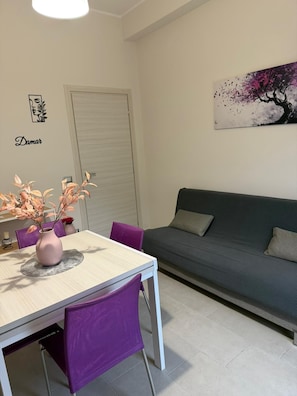 Ein moderner Wohnraum mit Esstisch, violetten Stühlen, einem grauen Sofa und einem an der Wand hängenden Kunstwerk.