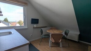 Een kleine, knusse kamer met schuine wanden, een ronde tafel met stoelen, een aan de muur bevestigde televisie en een raam met uitzicht op een woonwijk.