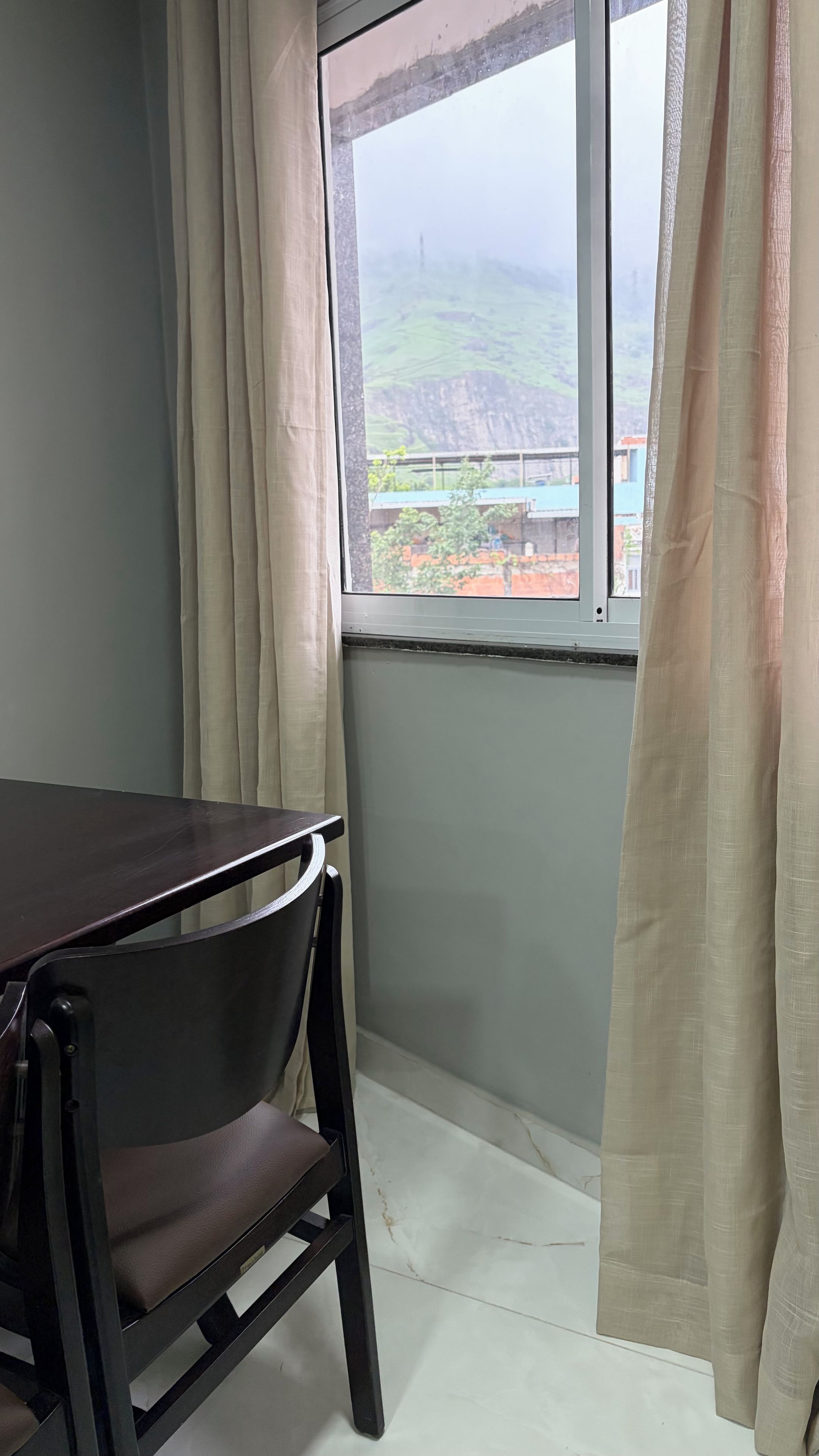 A room with a table and a chair, a window with curtains, and a view of a hilly landscape.