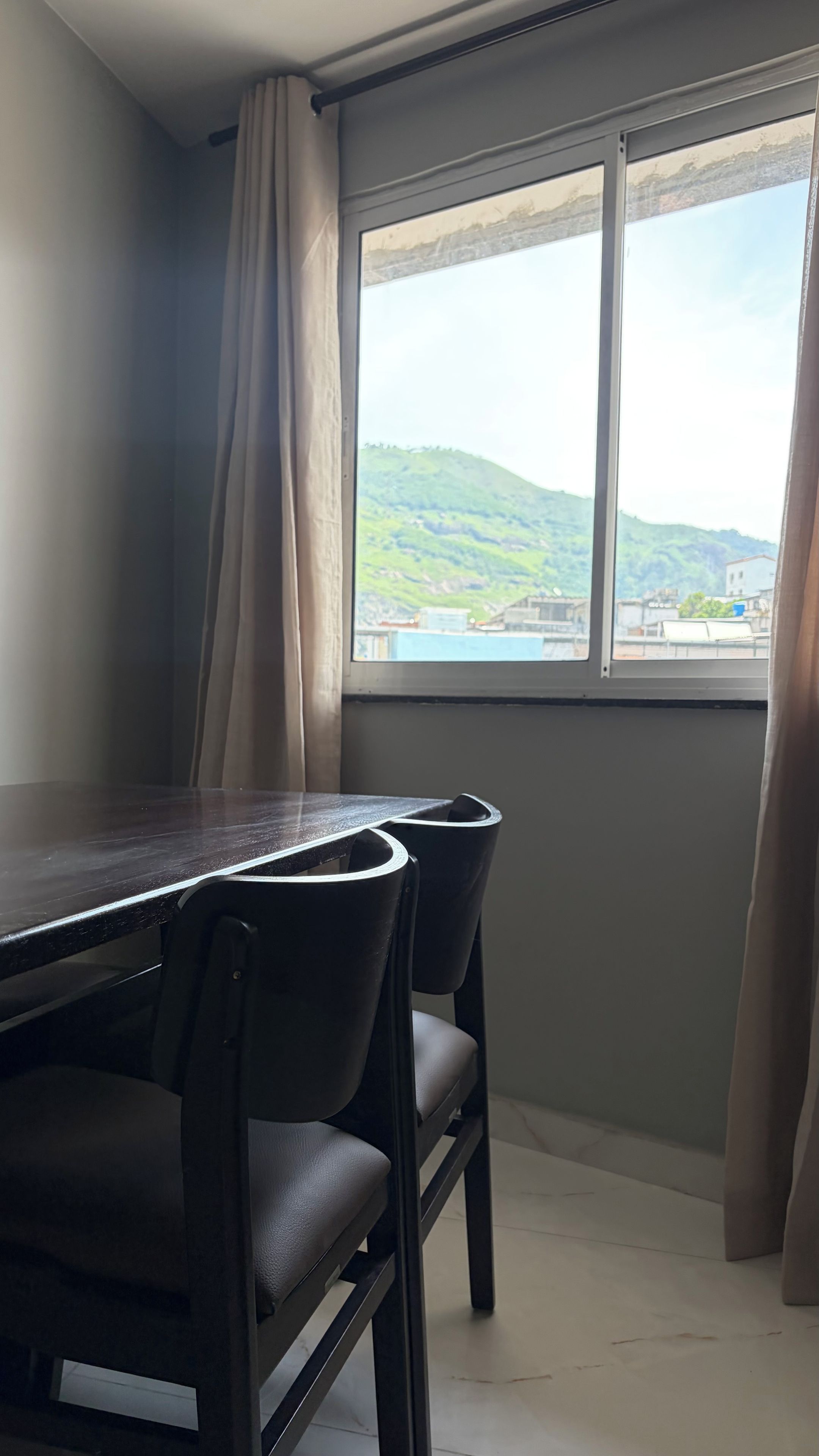 A room with a dining table and chairs, a window with a view of a green landscape, and a curtain partially drawn.