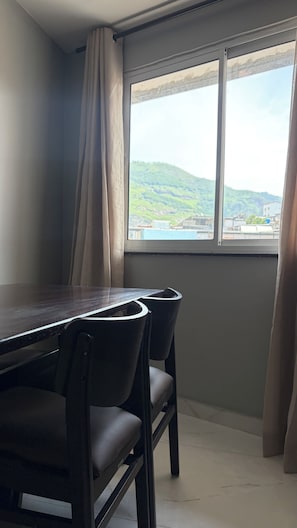 A room with a dining table and chairs, a window with a view of a green landscape, and a curtain partially drawn. - New Listing (Rio de Janeiro)