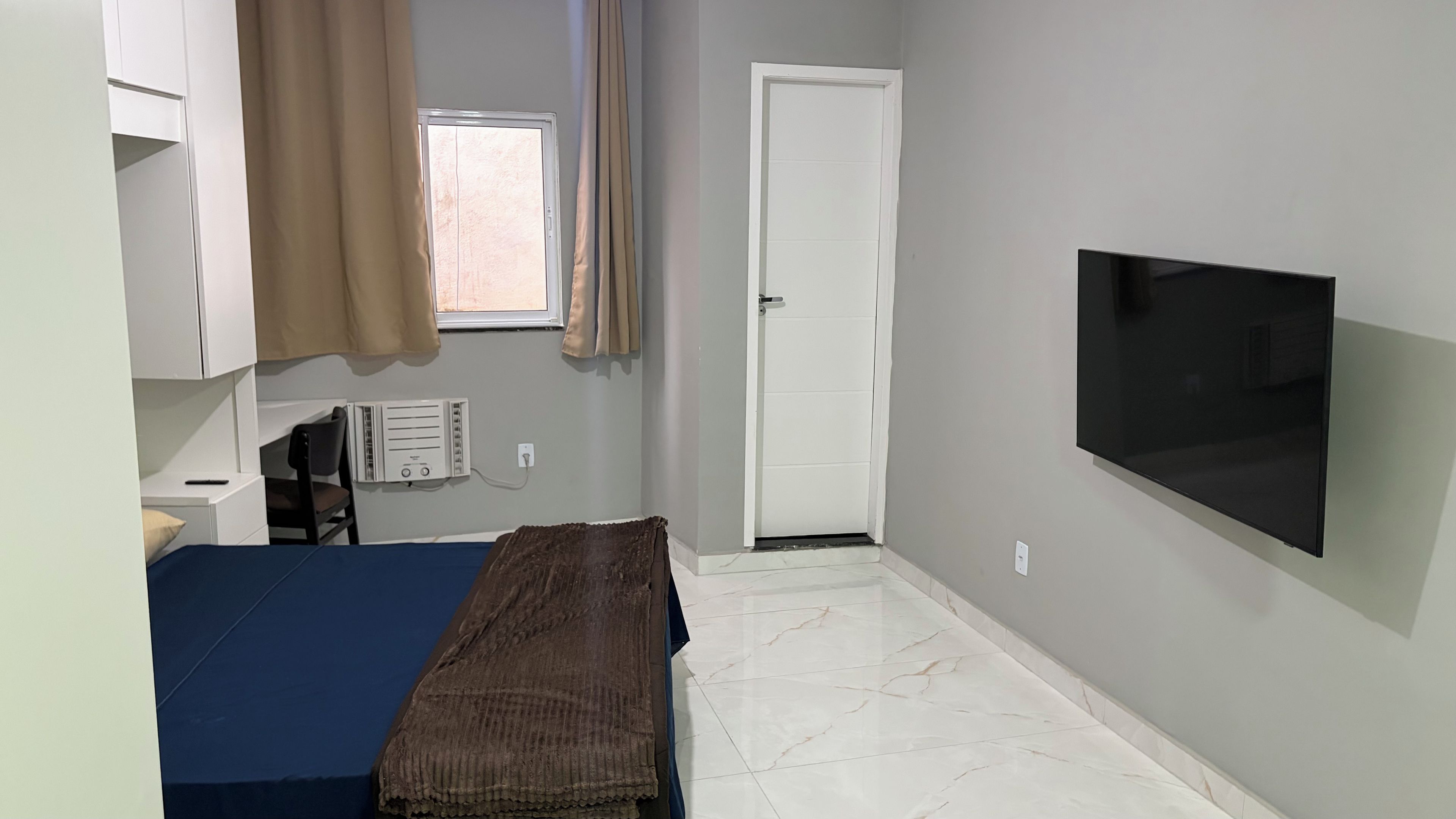 A bedroom with a bed, a TV, a window, a door, and a radiator.