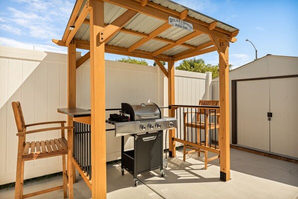 Een houten pergola met een barbecue en twee stoelen, een overdekt terras en een witte hek achterin.