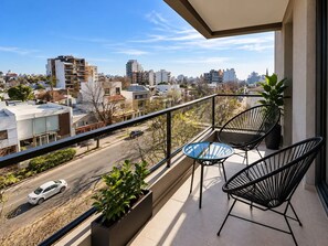 Un balcon avec deux chaises et une table, donnant sur une rue bordée d’immeubles et où l’on aperçoit une voiture.