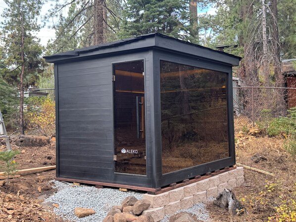 Una pequeña estructura de madera oscura con puerta de cristal, rodeada de un entorno natural con árboles y grava.
