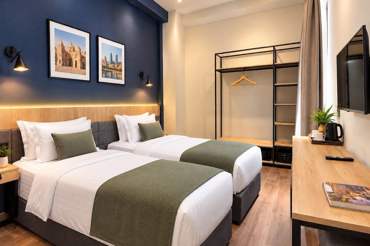 Quarto de hotel com duas camas, uma escrivaninha com cafeteira, televisão e guarda-roupa.