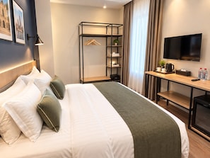 Quarto de hotel com cama, escrivaninha, televisão de tela plana, cabideiro e janela com cortinas.