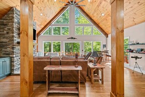 A spacious living room with a stone fireplace, a large wooden ceiling, and a view of the outdoors through large windows.