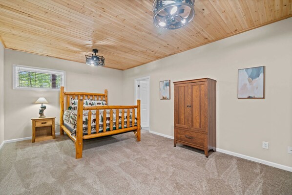 A bedroom with a wooden ceiling, a bed with a forest-themed bedspread, a nightstand with a lamp, a wooden wardrobe, and two framed pictures on the wall.