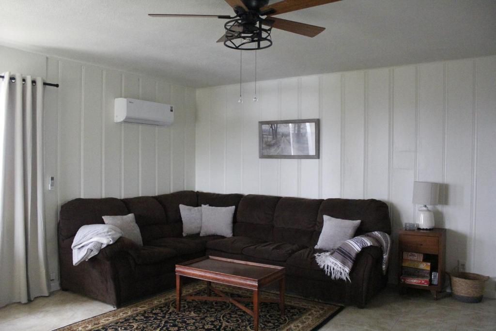Sala de estar com sofá seccional marrom escuro, mesa de centro de madeira, tapete estampado, ventilador de teto, aparelho de ar condicionado, quadro emoldurado, mesa lateral com abajur e estante de livros.