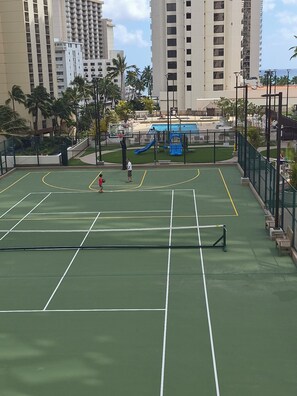 Un court de tennis en plein air, doté d’un filet, entouré d’une clôture, avec des palmiers en arrière-plan.