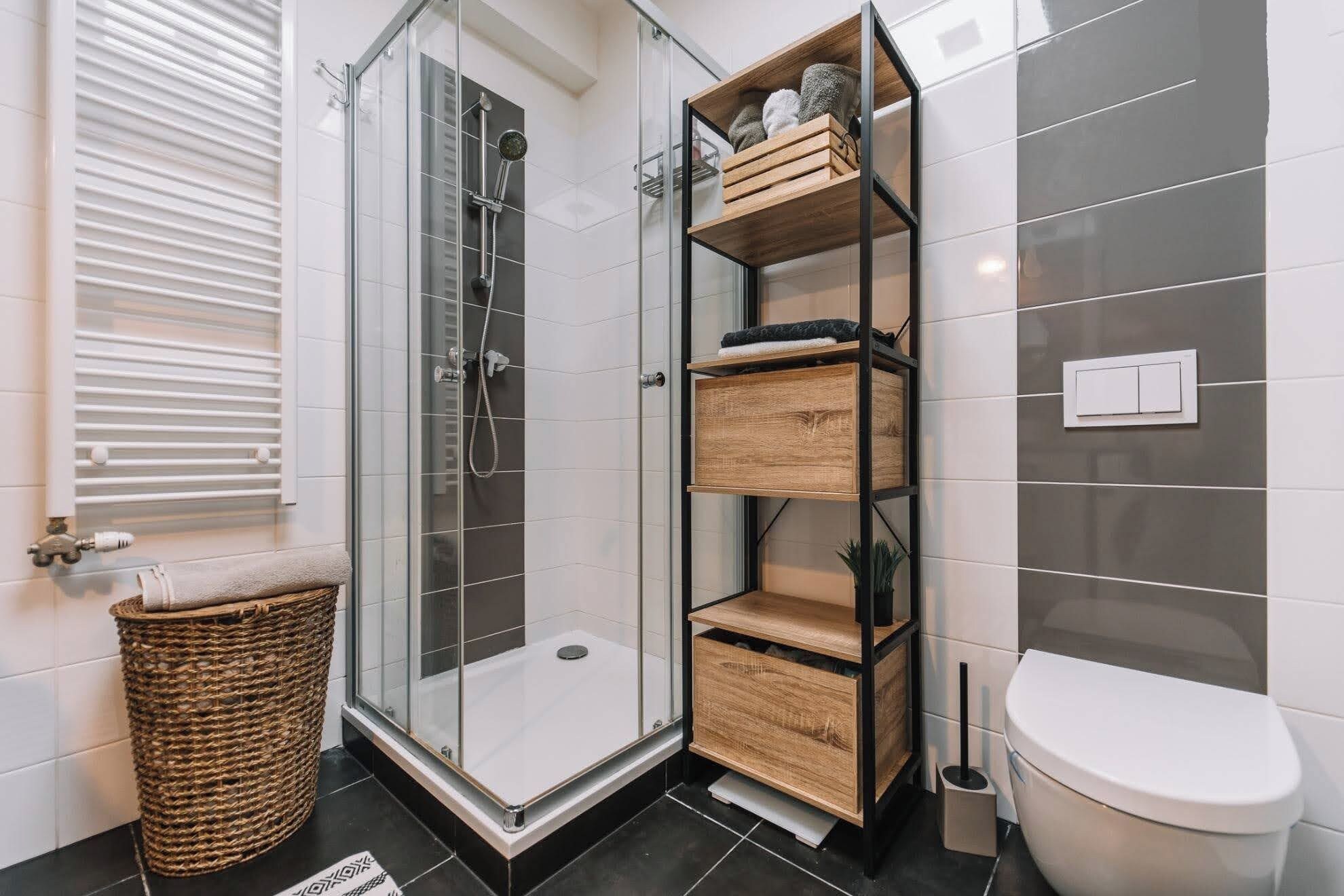 Et moderne badeværelse med glasbrusekabine, indbygget hyldeelement i træ, et hvidt toilet og en fletkurv.