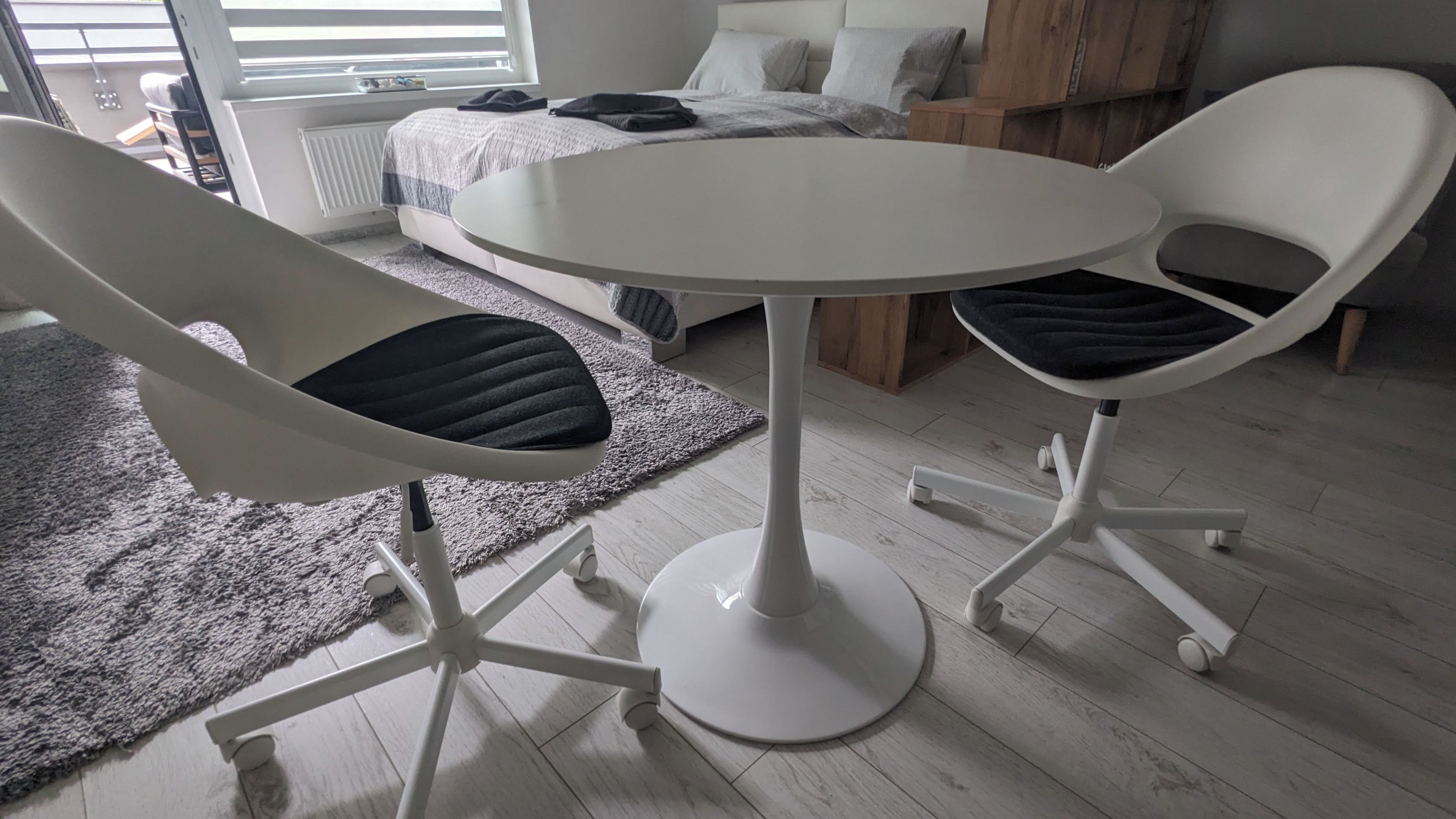 A modern bedroom with a round table and two chairs, a bed with a grey duvet, and a grey rug.