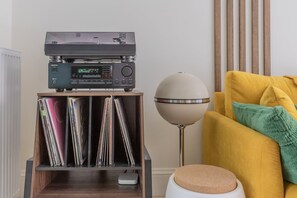 Huoneessa on puinen hylly, jossa on CD-levyjä, musta äänentoistojärjestelmä, pallomainen valaisin ja keltainen sohva, jonka päällä on vihreä koristetyyny.