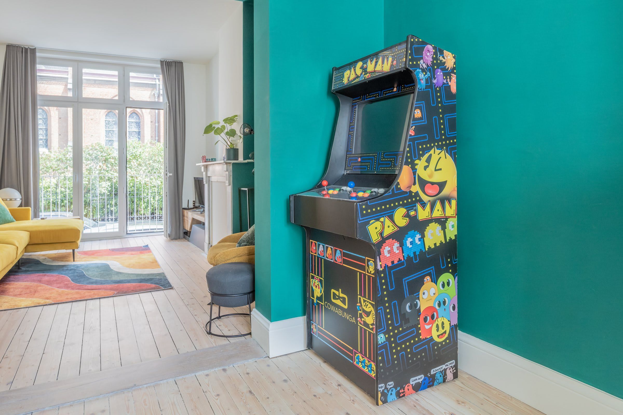 Huoneessa on turkoosinsävyinen korosteseinä, Pac-Man-peliautomaatti, keltainen sohva ja suuri ikkuna, josta avautuu näkymä vehreään maisemaan.