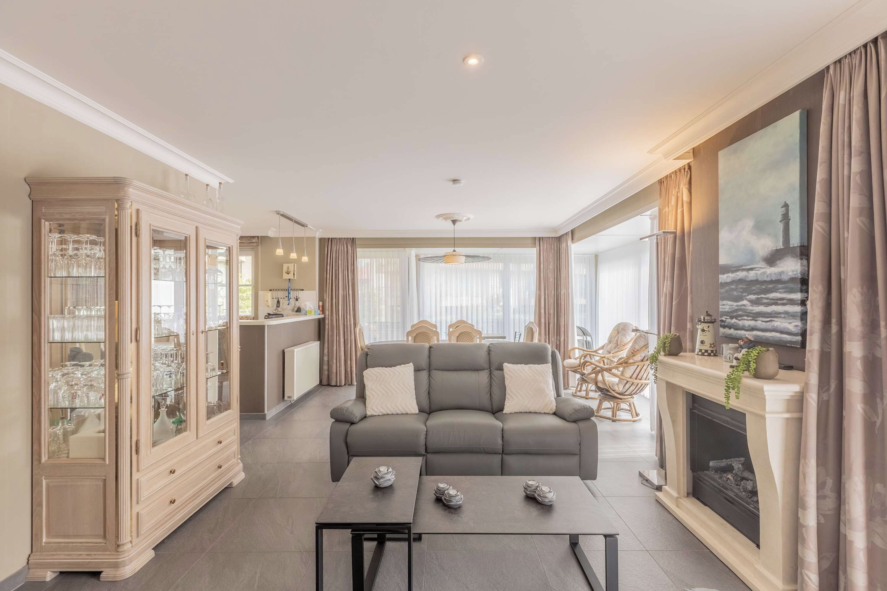 Een woonkamer met een grijze bank, een eethoek, een keuken en een open haard, inclusief een schilderij van een vuurtoren.