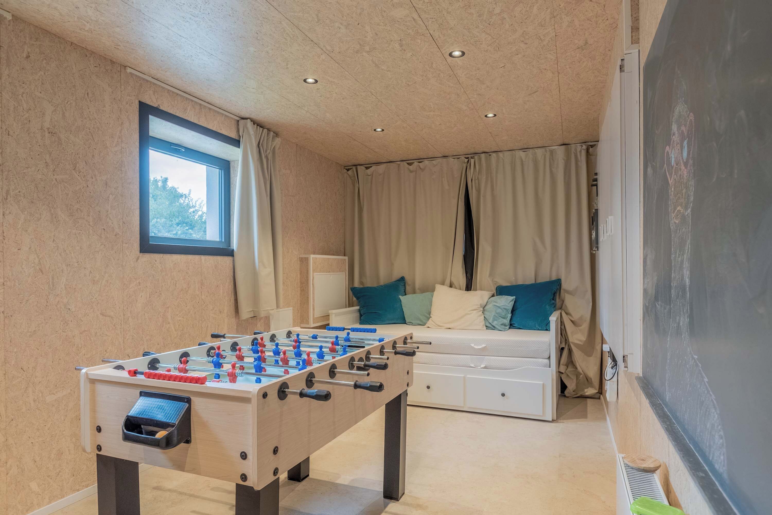 Ein Zimmer mit Kicker, einem Bett mit Kissen und einem Fenster.