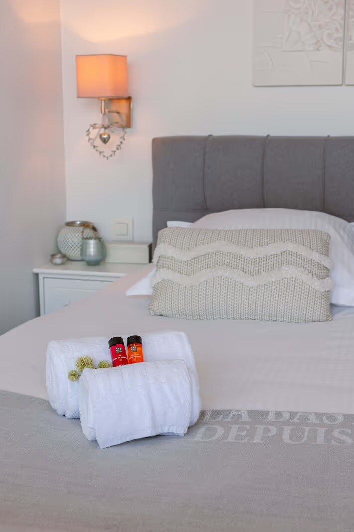 A neatly made bed with white linens, a grey headboard, and two pillows. There is a bedside table with a lamp and a decorative item.