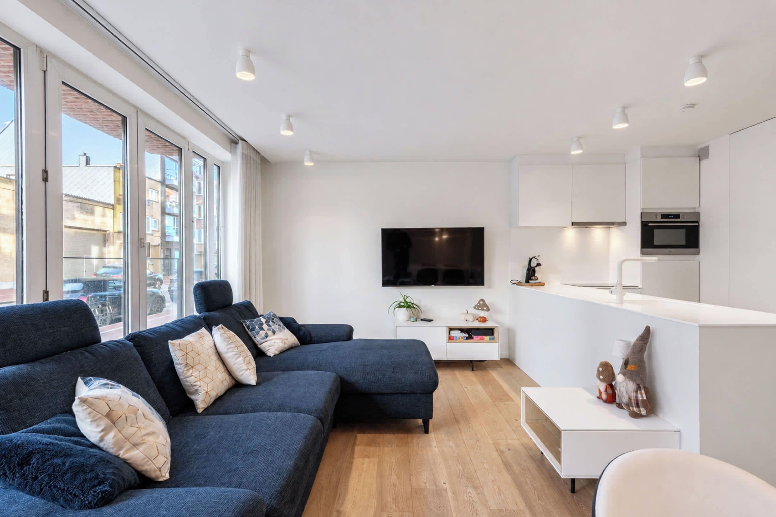 A modern living room with a dark blue sofa, a flat-screen TV, and a minimalist kitchen area.