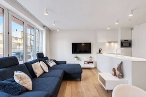 A modern living room with a dark blue sofa, a flat-screen TV, and a minimalist kitchen area. - La Mer Moiselle · 2 bedrooms & private parking (Knokke-Heist)