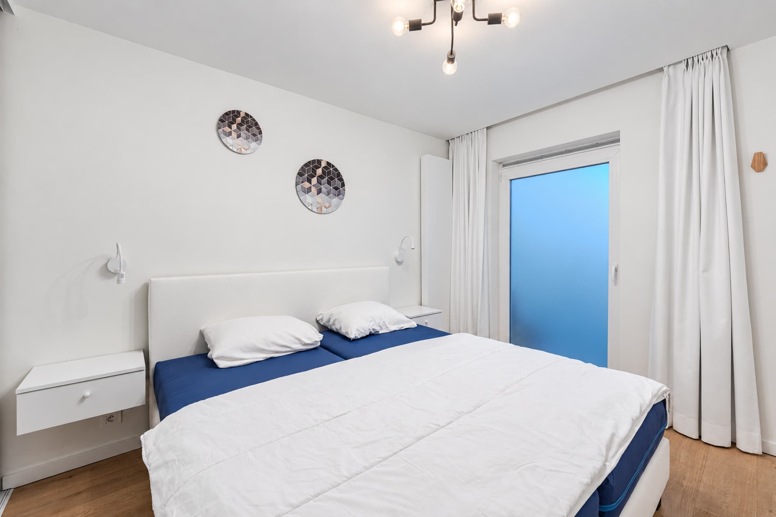 A bedroom with a bed, a nightstand, a wall-mounted light fixture, and a door with curtains.