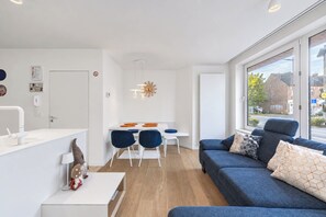 Una sala moderna con un sofá azul, un comedor con mesa blanca y sillas azules, y un amplio ventanal con vista a la calle.