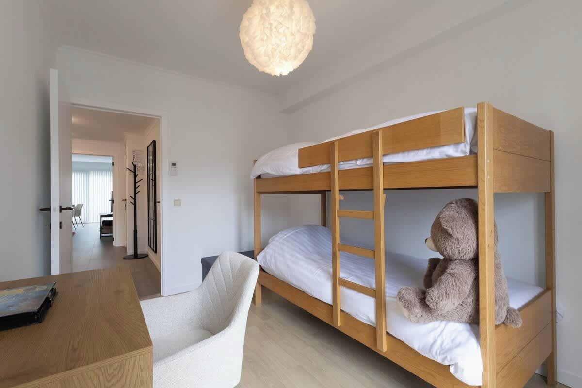 A bunk bed with a teddy bear on the lower bunk, a desk with a chair, and a hanging light fixture.