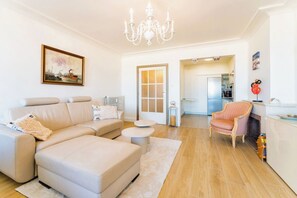 Ein Wohnzimmer mit einer Couch, Hocker und einem Sessel. Es gibt eine Chandelüster, ein Gemälde an der Wand und einen Blick in die Küche.