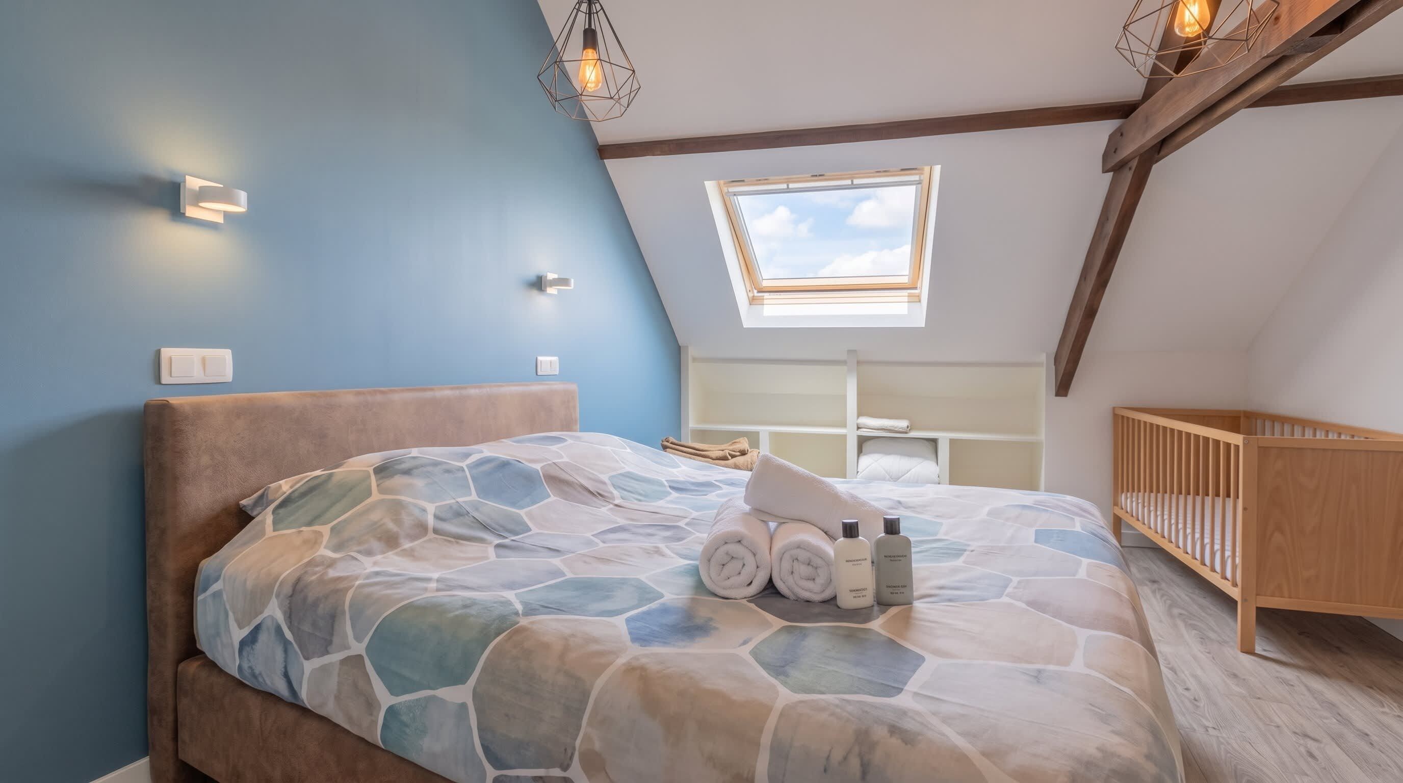 Ein Schlafzimmer mit schrägem Dach, einem Bett mit gemustertem Überwurf, einem Kinderbett und einem Dachfenster.